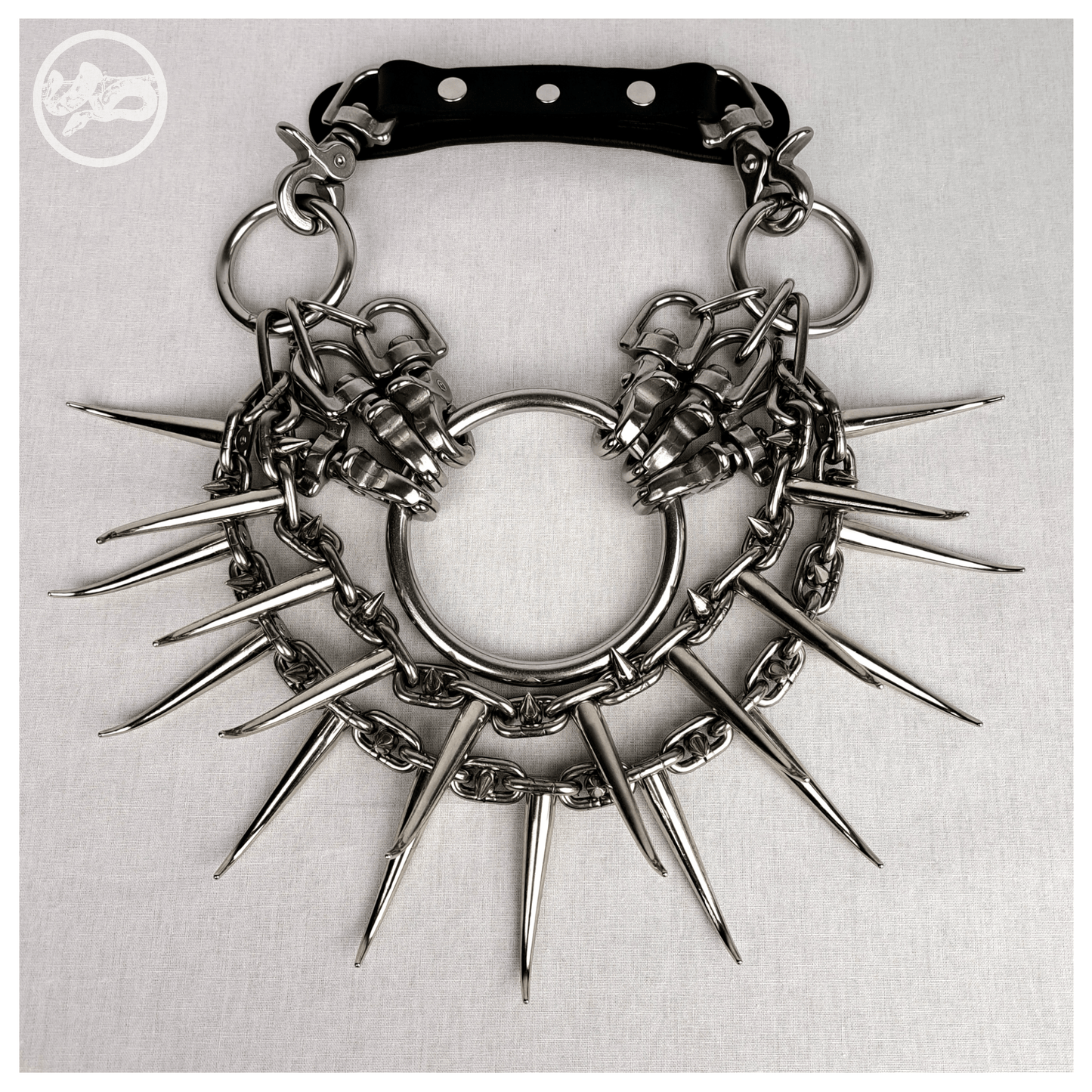 Brutalist necklace FEGEFEUER laid out flat in a front view, featuring studded stainless steel chains with long zinc alloy claw spikes, a large central O-ring, and a reclaimed leather neckpiece with stainless steel carbines, by skinpressedtight.