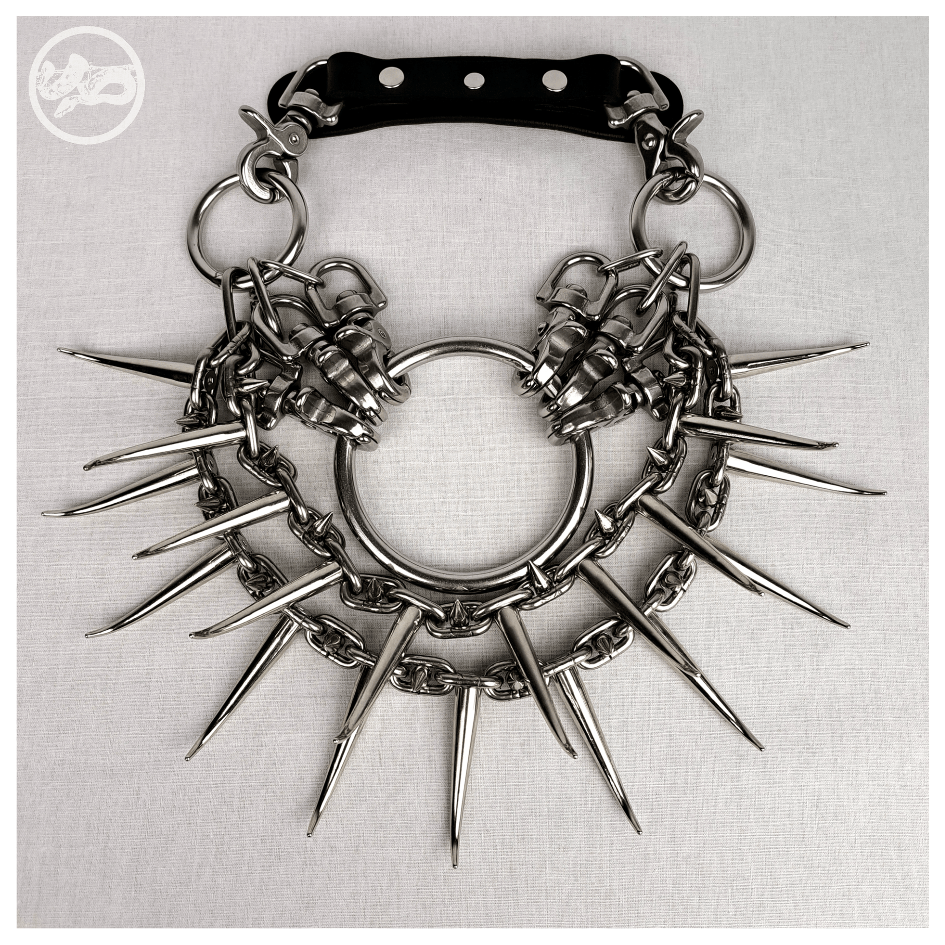 Brutalist necklace FEGEFEUER laid out flat in a front view, featuring studded stainless steel chains with long zinc alloy claw spikes, a large central O-ring, and a reclaimed leather neckpiece with stainless steel carbines, by skinpressedtight.