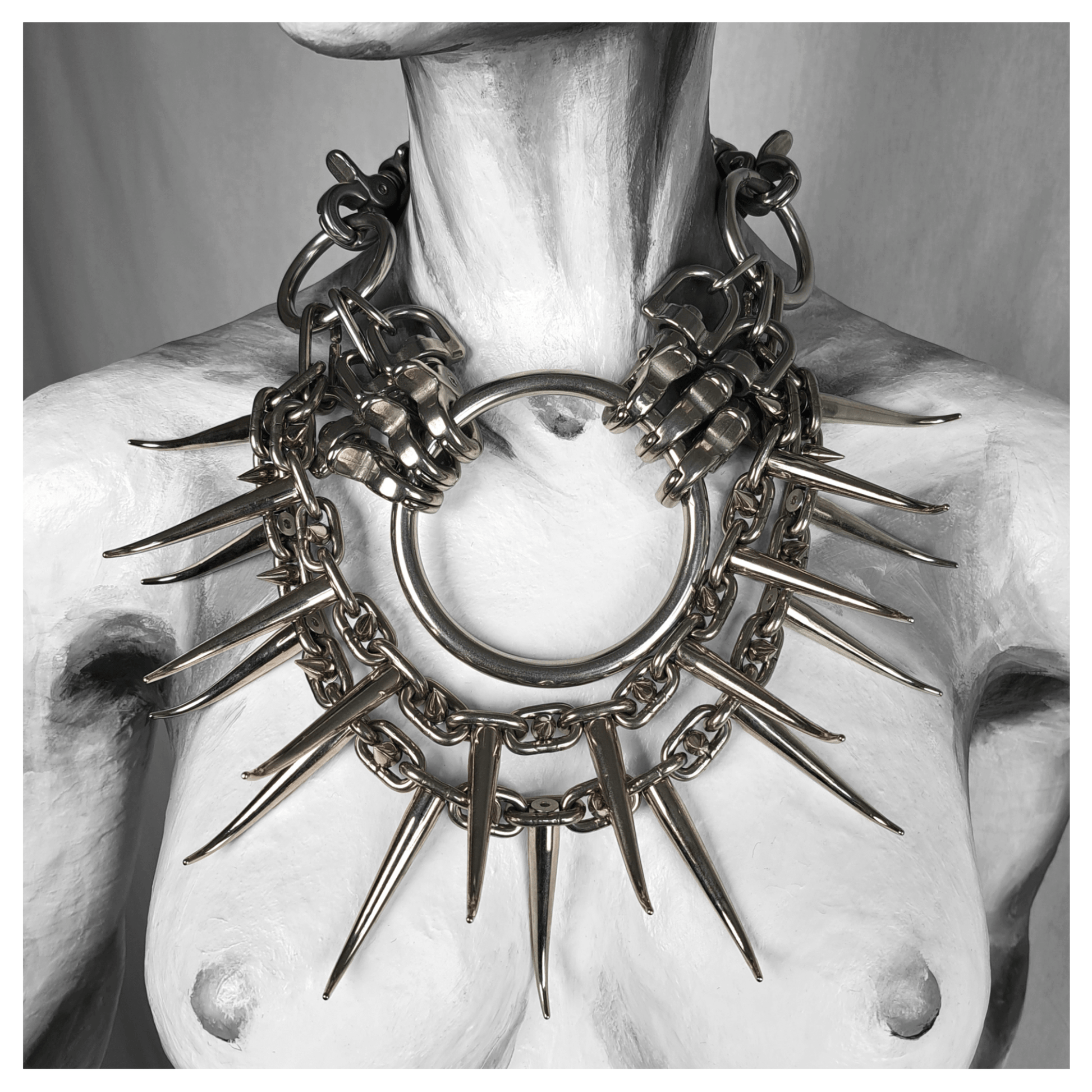 Brutalist necklace titled FEGEFEUER displayed on a mannequin, featuring studded stainless steel chains with long zinc alloy claw spikes, a large central O-ring clasp, and a reclaimed leather neckpiece closed with carbines, by skinpressedtight.