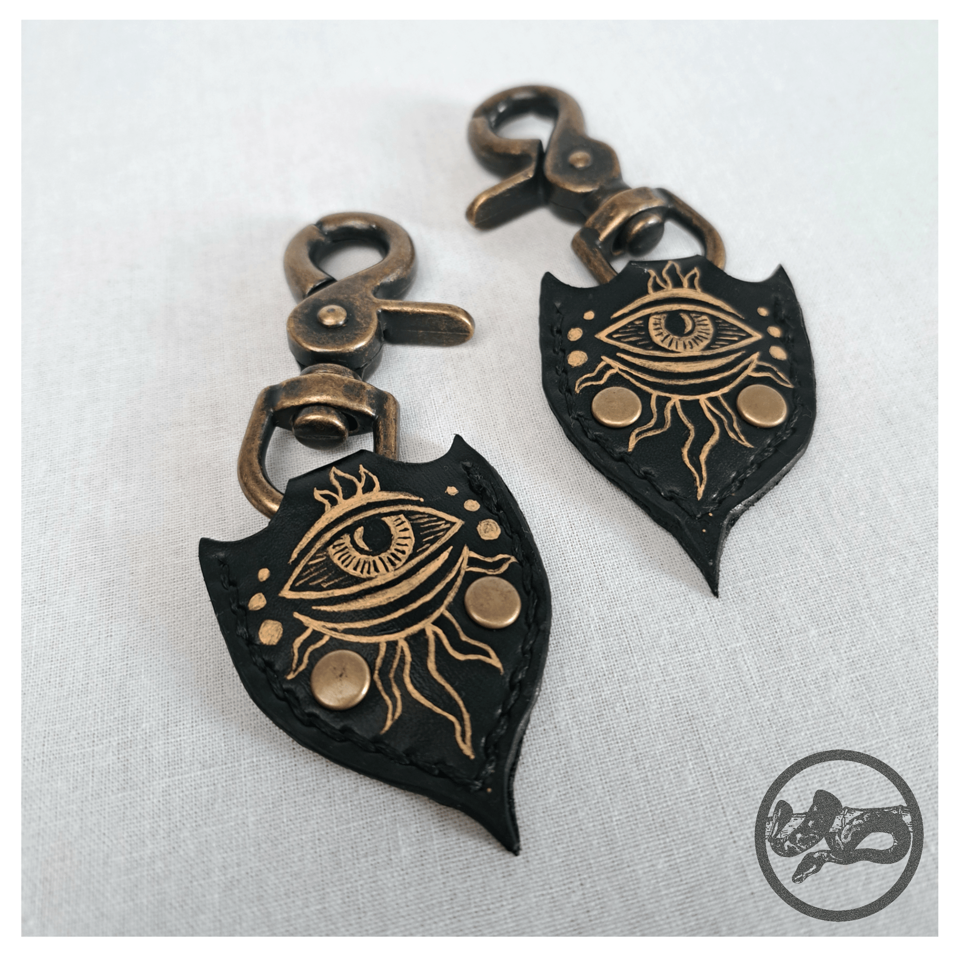 Detail view of JOTKAELL spearhead-shaped earrings and ear charms on white background, highlighting leather pieces with handpainted golden eyes and tentacle ornaments, attached with brass carbines — gothic handcrafted accessories by skinpressedtight.