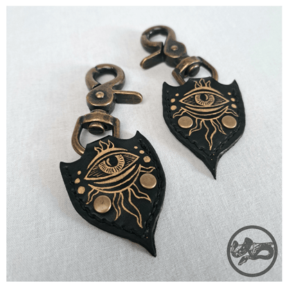 Detail view of JOTKAELL spearhead-shaped earrings and ear charms on white background, highlighting leather pieces with handpainted golden eyes and tentacle ornaments, attached with brass carbines — gothic handcrafted accessories by skinpressedtight.