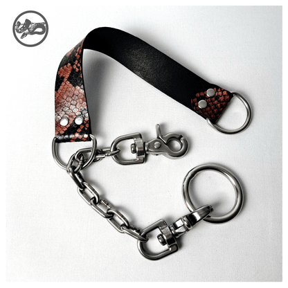 BOAKAPITEL – I. RED PYTHON choker open on white: one scissor carabiner unclipped from the O-ring, showing the chain running through the D-ring of the python-print leather band. Handmade by skinpressedtight
