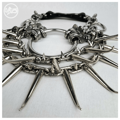 Detail of the FEGEFEUER necklace laid flat: polished scissor carabiners gripping the large O-ring, heavy stainless steel chain studded with long horn spikes and small cone spikes. Handmade by skinpressedtight