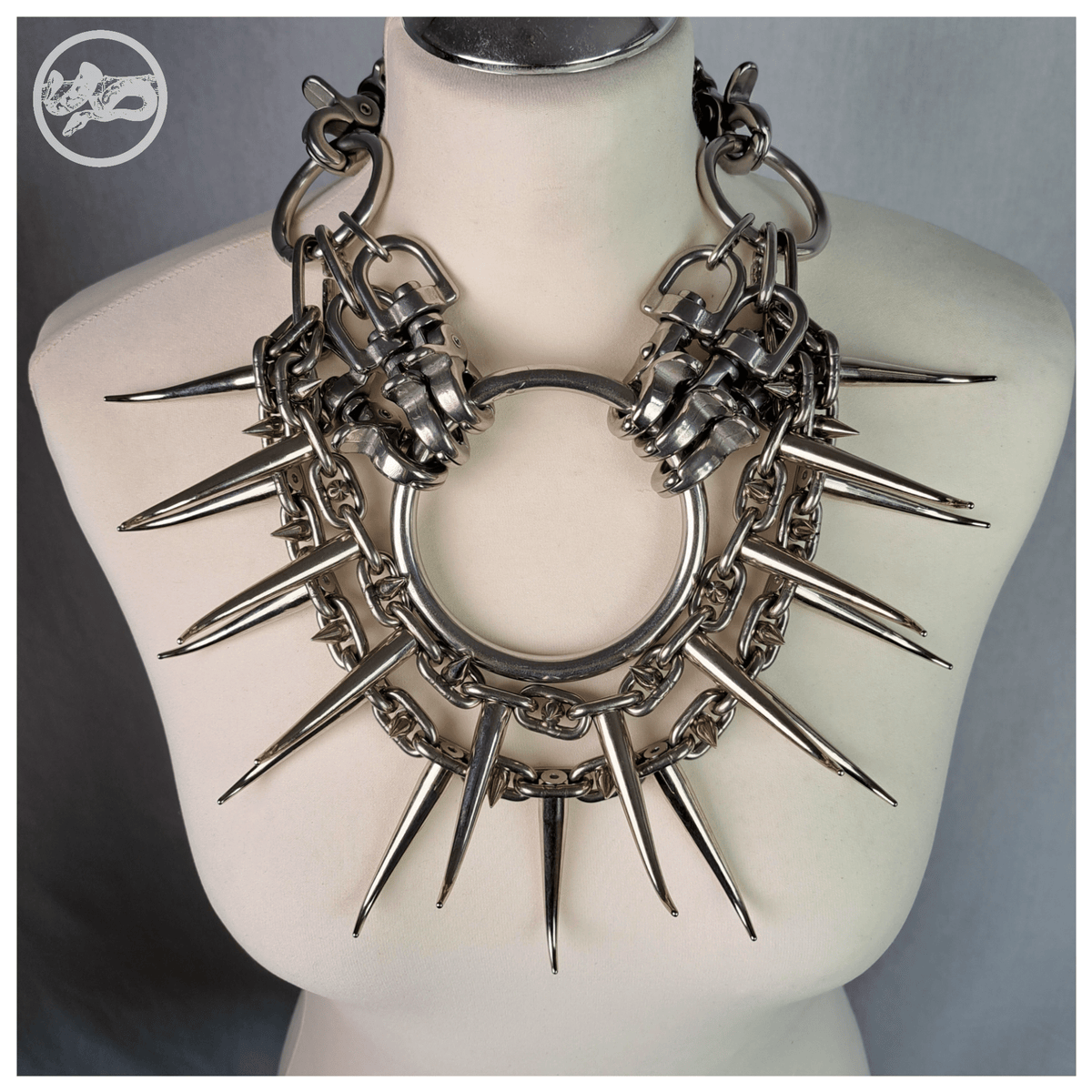 FEGEFEUER necklace on a white dress form, front view: a heavy all-metal chest piece with a central O-ring, carabiner clusters and two rows of long spikes fanning out over the chest like rays. Brutalist jewelry by skinpressedtight