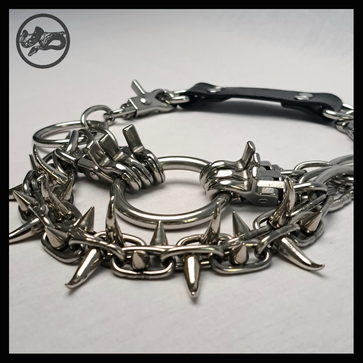 FEGEFLAMME necklace detail, angled on the table: polished scissor snap hooks stacked beside the 40 mm ring, chain swags bristling with cone spikes and curved claw studs. Handmade by skinpressedtight