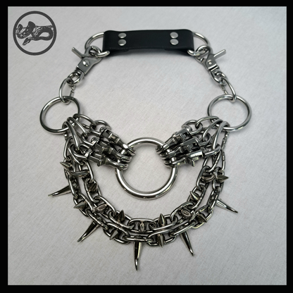 FEGEFLAMME necklace closed, laid flat: black leather back strap with D-rings at the top, side O-rings, snap-hook clusters and the spiked chain swags around the large O-ring. Handmade by skinpressedtight