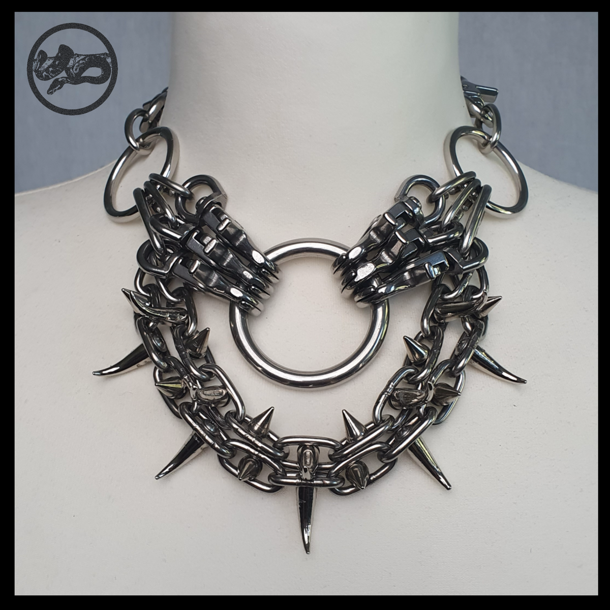 FEGEFLAMME spiked chain necklace on a cream dress-form bust, front view: centre ring flanked by snap-hook clusters, two U-shaped swags of spiked chain on the collarbones. Handmade by skinpressedtight