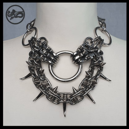 FEGEFLAMME spiked chain necklace on a cream dress-form bust, front view: centre ring flanked by snap-hook clusters, two U-shaped swags of spiked chain on the collarbones. Handmade by skinpressedtight