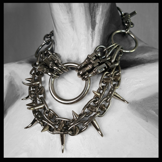 FEGEFLAMME statement necklace worn on a painted mannequin, black and white: a large stainless O-ring between two clusters of snap hooks, with two swags of chain set with spikes and claw studs below. Handmade by skinpressedtight