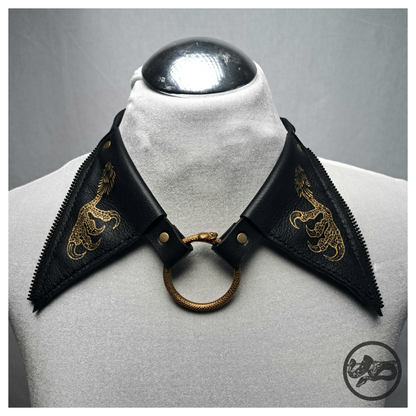 FLATTERTIER – BABA JAGA leather collar necklace on a white dress form, front: wide pointed black flaps painted with gold chicken feet, brass snake ring at the centre. Handmade by skinpressedtight