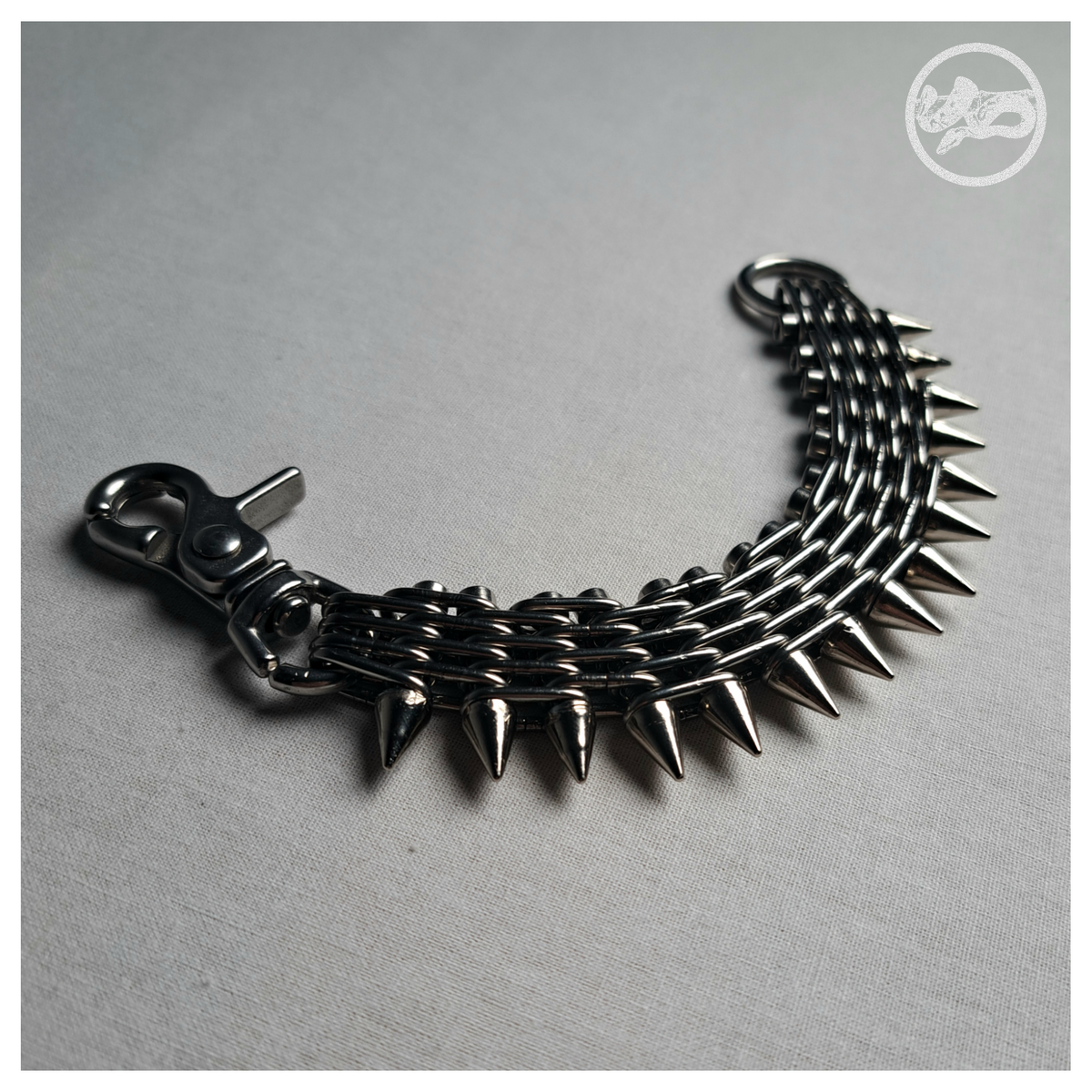 GEWALTBEREIT spiked chain bracelet open, lying in an arc: the stacked stainless chain band, cone spikes along the outer edge, snap hook at one end and O-ring at the other. Handmade by skinpressedtight