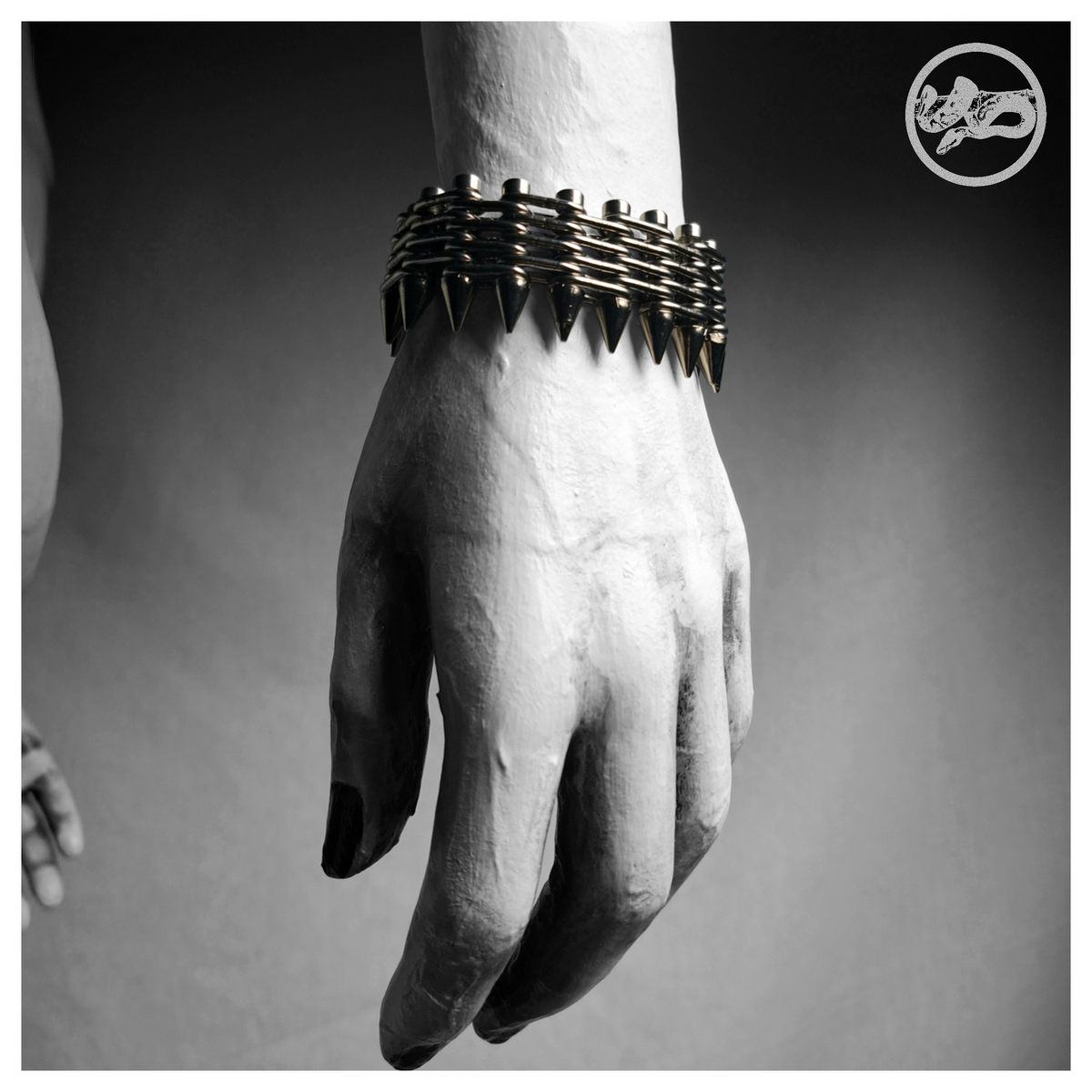 GEWALTBEREIT spiked bracelet worn on a painted mannequin's wrist, black and white: a flat band of stacked stainless chain on screws with a row of cone spikes along one edge. Handmade by skinpressedtight
