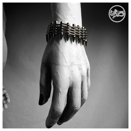 GEWALTBEREIT spiked bracelet worn on a painted mannequin's wrist, black and white: a flat band of stacked stainless chain on screws with a row of cone spikes along one edge. Handmade by skinpressedtight