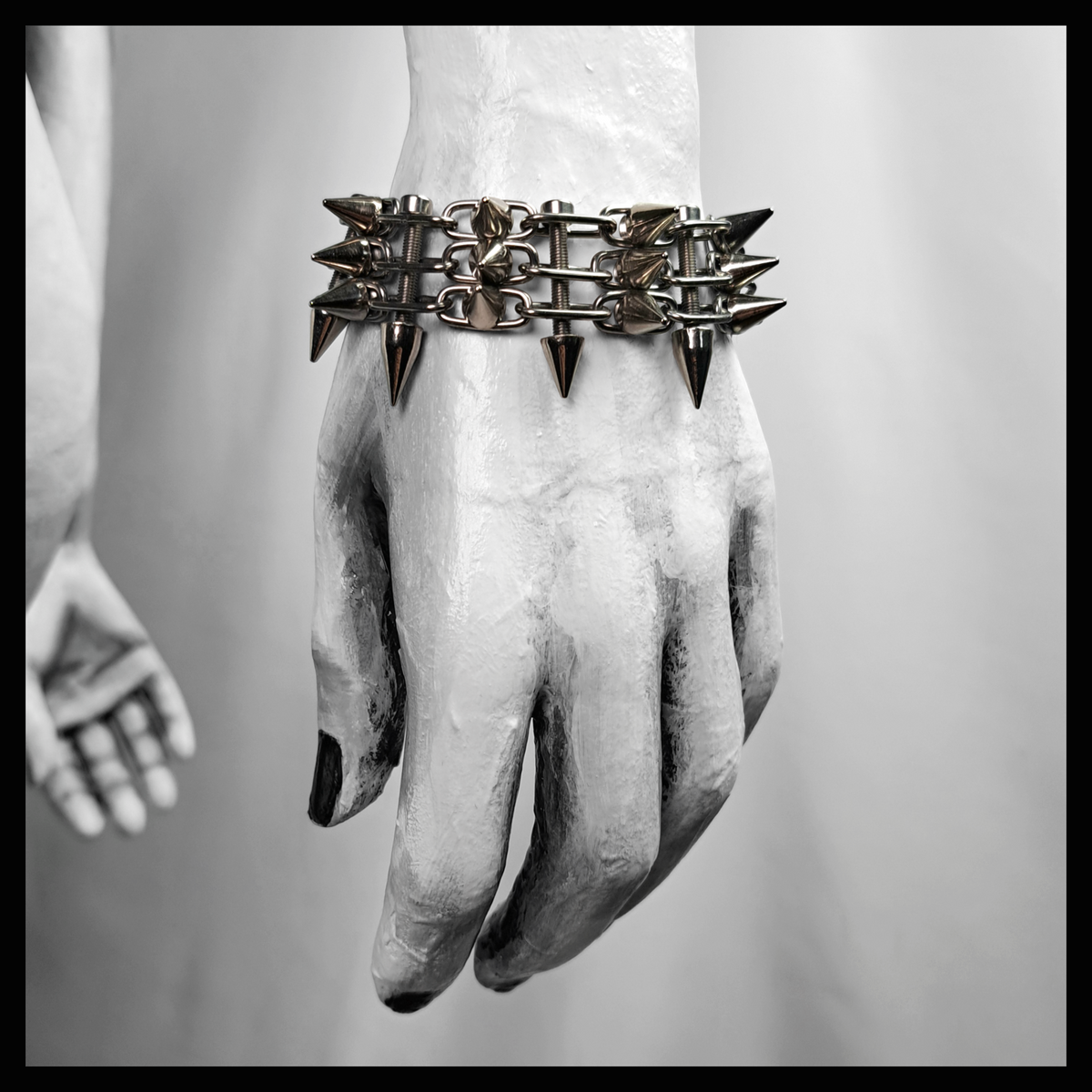 SCHALTKREIS spiked bracelet worn on a painted mannequin's wrist, black and white: a ladder band of fine stainless steel chain with rows of small cone spikes. Handmade by skinpressedtight