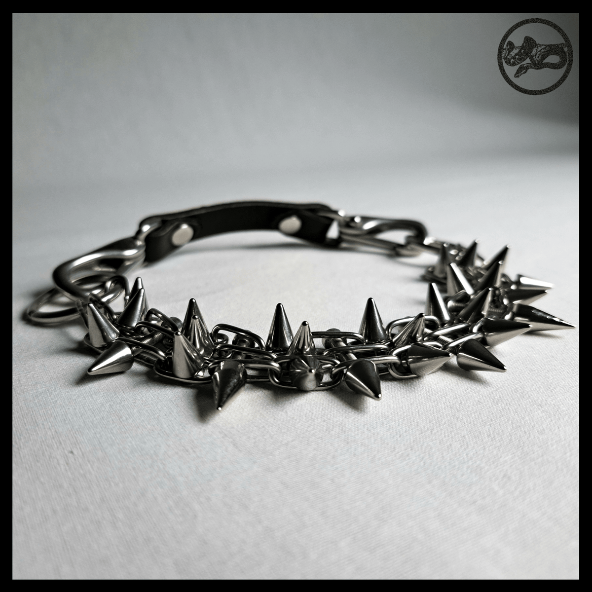 STŌRENFRIED spiked chain choker closed on a table, low angle: cone spikes along the band, black leather strap at the back. Handmade by skinpressedtight