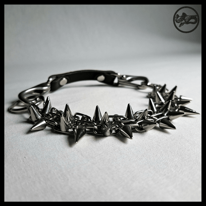 STŌRENFRIED spiked chain choker closed on a table, low angle: cone spikes along the band, black leather strap at the back. Handmade by skinpressedtight